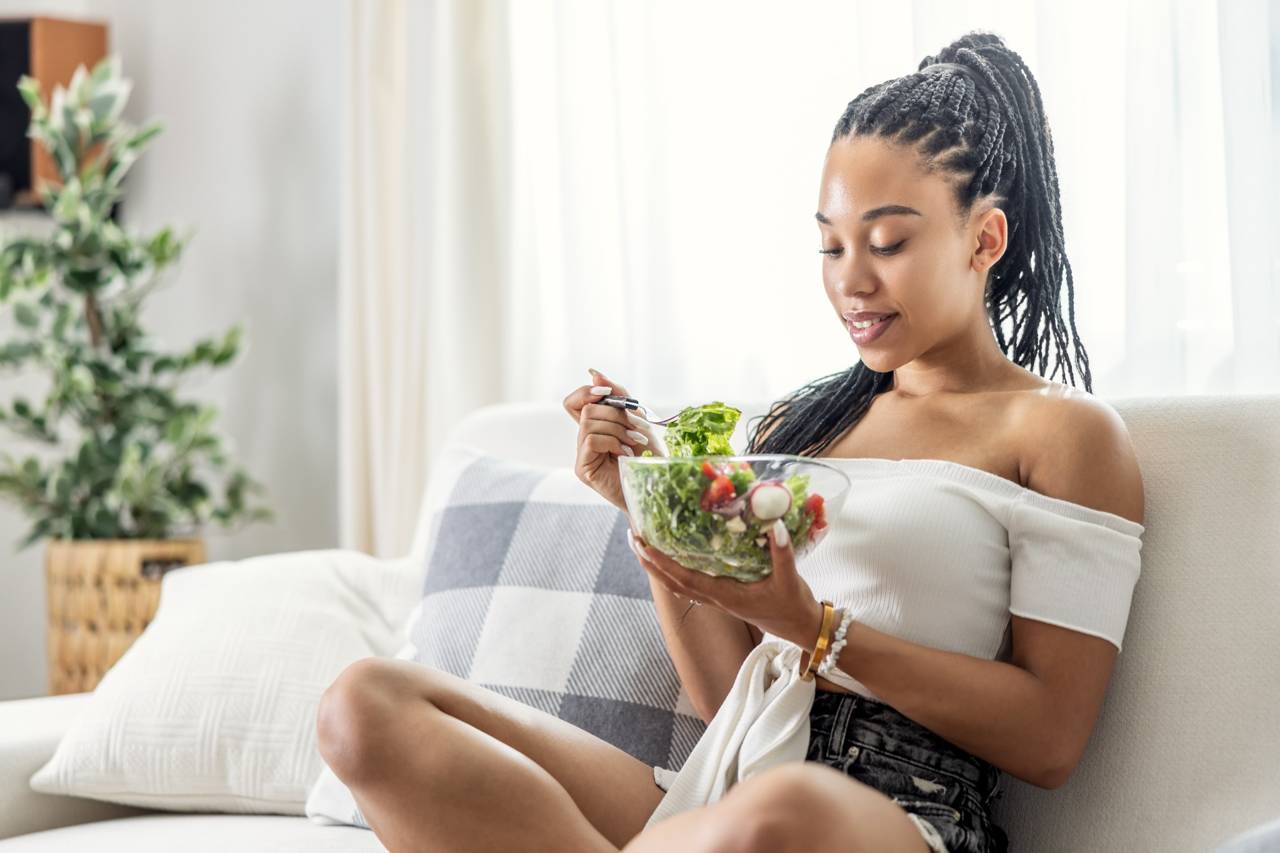 Young woman sitting on a couch, enjoying a fresh salad in a bright living room.