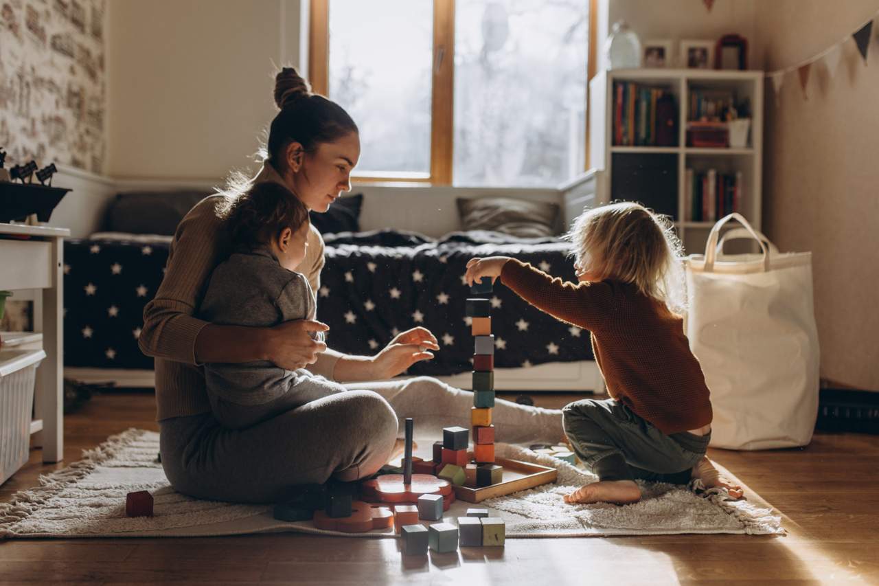 Mother sits on floor holding toddler while older sibling builds tall tower of colorful blocks in sunlit bedroom.
