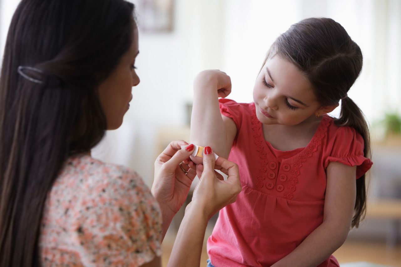Mother applying bandage to young girl’s elbow as the child lifts her arm to look.