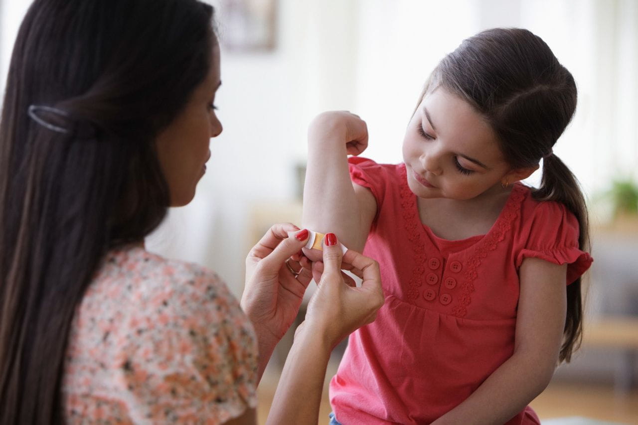 Mother applying bandage to young girl’s elbow as the child lifts her arm to look.