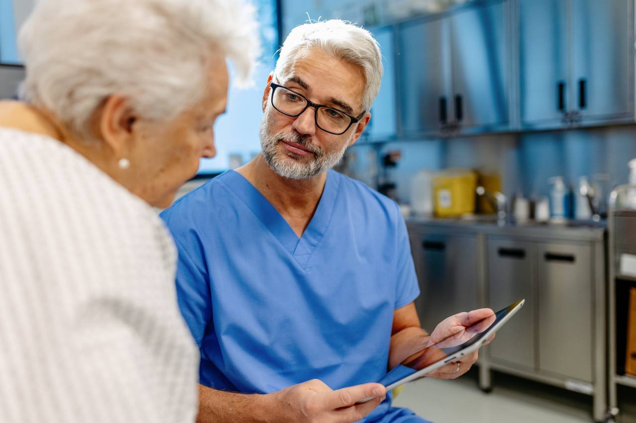 Doctor in blue scrubs holding a tablet while talking to an elderly patient.