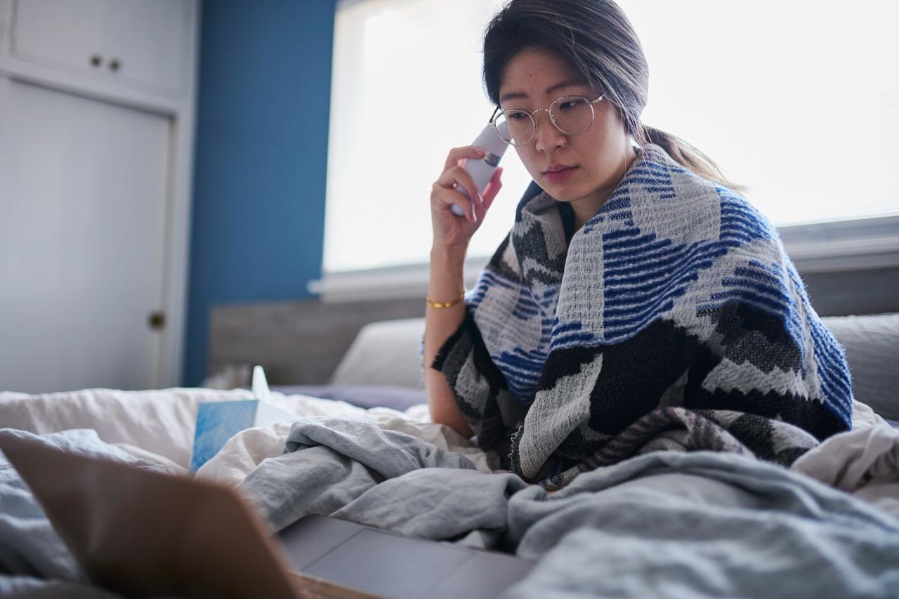 Woman wrapped in patterned blanket checks temperature with digital thermometer while working on laptop in bed.