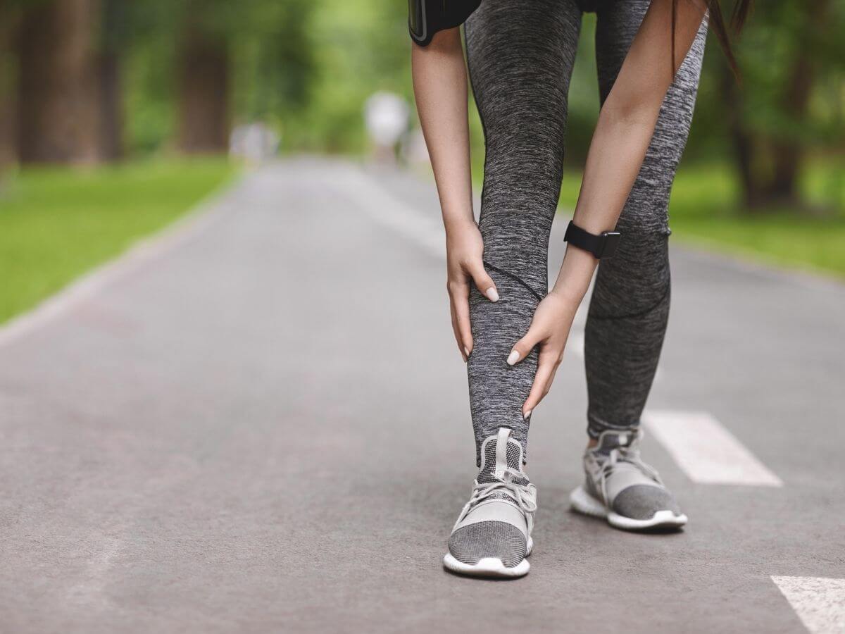 Photo of a woman running outside from the legs down. She is bent over holding her shin in pain.